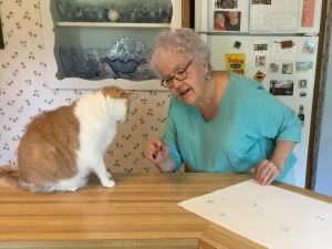 Artist Jo Myers-Walker in conversation with an orange-and-white cat. Jo is holding a paintbrush and standing next to paper with a faint line of paw prints.